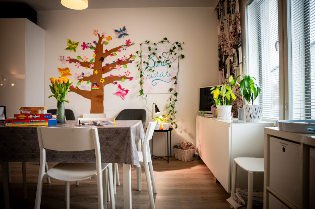 Provesta Korten tiloista kuva, jossa on ruokapöytä harmaalla pöytäliinalla ja kaksi vakoista tuolia. Pöydän pääll on keltaisia tulppaaneita ja lautapelejä. Oikealla puolella huonetta on ikkuna joka tuo valoa huoneesee. Ikkunan edessä vakoinen lipasto, jonka päällä kasveja ja televisio. Huoneen takana olevassa seinässä on värikäs koristeellinen puu sekä taulu, jossa lukee "Sinä riität".