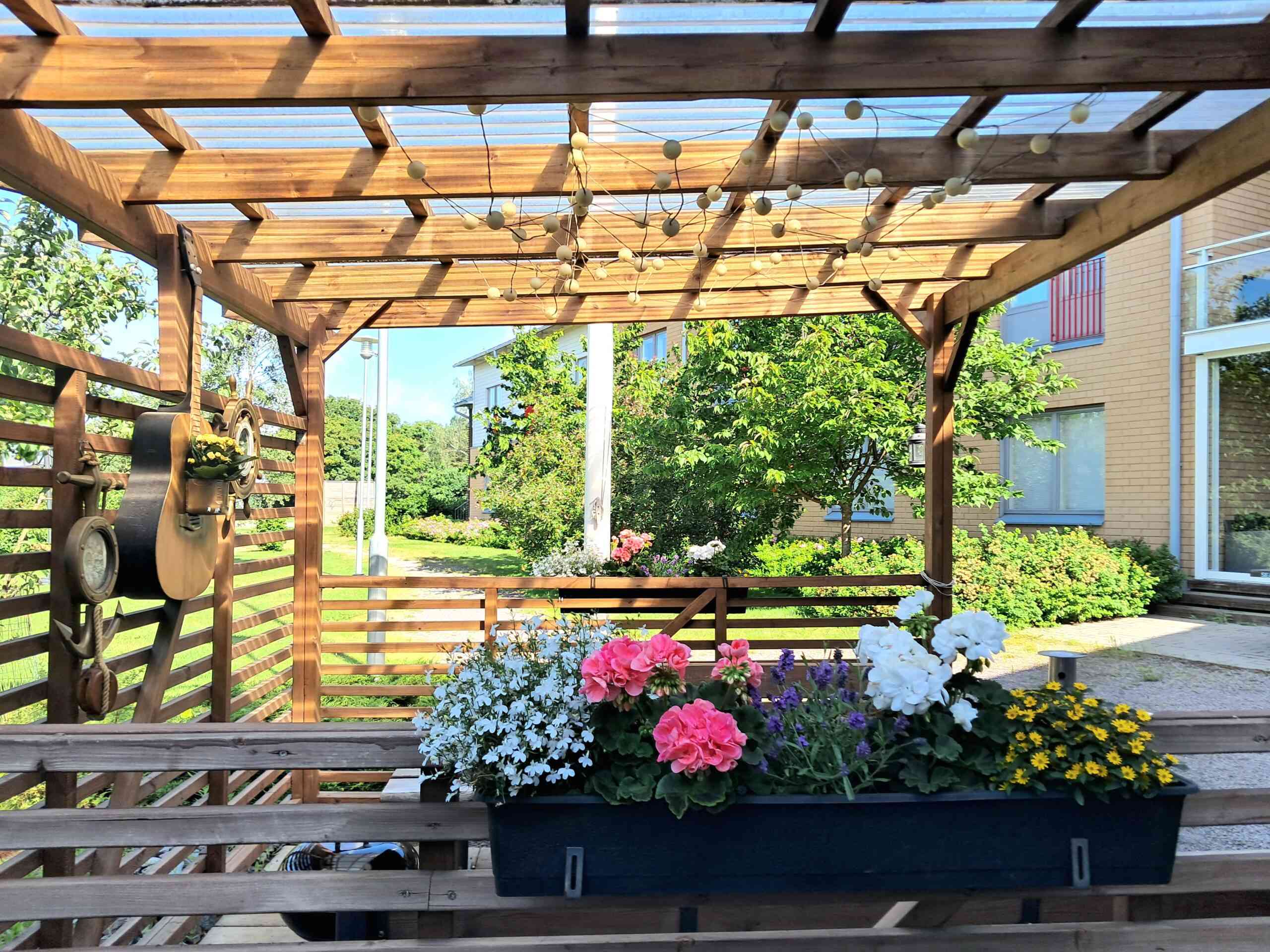 Yksikön pihalta otettu kuva, jossa puinen katos ja kukka-asetelma